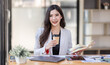 © David - Asian Businesswoman having a tea, sitting on desk in workplace, writing down notes, opened laptop in document the financial report, business plan investment, finance analysis concept.