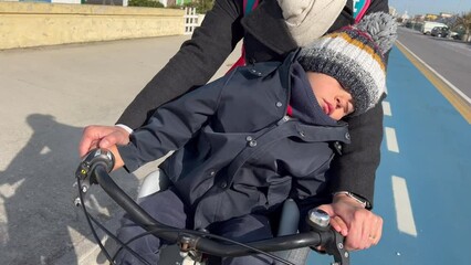 Wall Mural - Child sleeping while riding bicycle with mother. One small kid napping seated in front of bike chair with parent in cyclist lane in street during autumn season