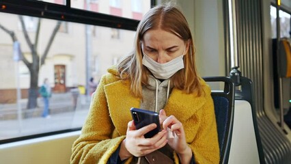Wall Mural - Public Transportation. Blonde woman in medical mask holding and using smartphone in a city bus. Female passenger looking away at window, waiting for her stop, browsing route. 