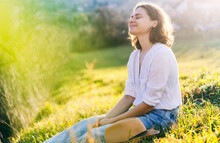Happy Pretty Young Cheerful Woman In White Shirt Sitting On Grass Enjoying Sunny Spring Day With Eyes Closed
