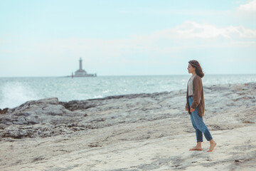 Wall Mural - woman walking by rocky sea beach in wet jeans lighthouse on background