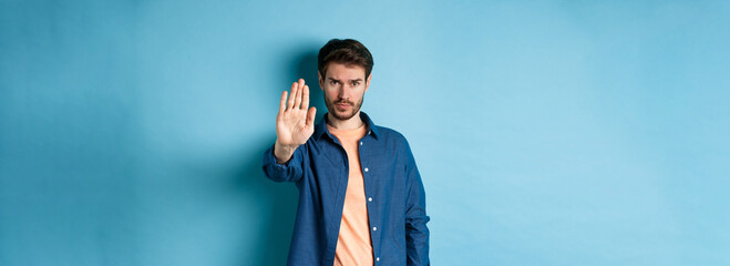 Upset man frowning and asking to stop, stretch out hand to prohibit or disagree with something bad, standing on blue background