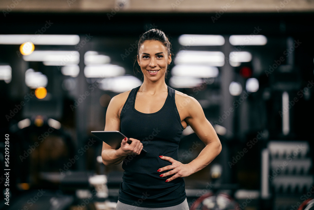 A fit muscular female personal trainer is holding tablet in her hands and smiling at the camera in a gym.