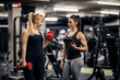 © dusanpetkovic1 - A female bodybuilder is lifting dumbbells in a gym while her female personal trainer is looking at her progress.