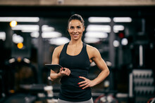 A Fit Muscular Female Personal Trainer Is Holding Tablet In Her Hands And Smiling At The Camera In A Gym.