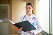 © Suriyo - Woman doctor holding folder in hospital background medicine, profession and healthcare concept - happy smiling female doctor in white coat with folder and stethoscope over hospital background