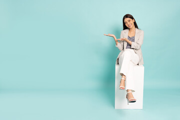 Happy young Asian businesswoman presenting or showing open hand palm with copy space for product and sitting on white box isolated on green background