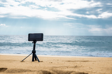  A smartphone camera on a tripod in the foreground stands on the seashore in cloudy weather. High quality photo