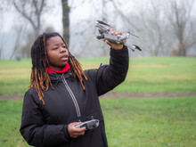Young african girl standing checking flying drone camera outdoors in a park in cold weather.