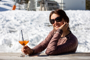 Woman, sitting on a table in the mountains, drinking aperol while having pause from skiing