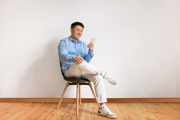 Wall Mural - Excited middle aged asian man using smartphone, networking in social media, sitting on chair over white studio wall