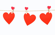 Three Red Hearts Are Attached To A Rope With Clothespins On A White Background. Beautiful Decor In The Form Of Hearts Or Decoration For Valentine's Day. Three Red Hearts Made Of Wood
