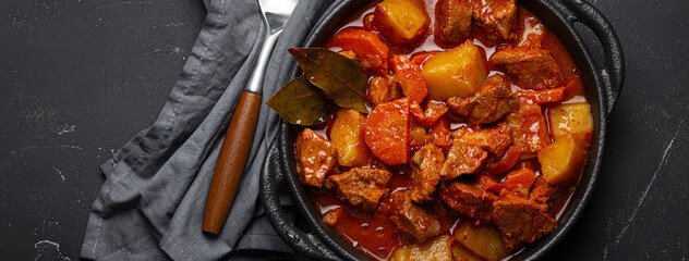 Beef meat stew with potatoes, carrot and delicious gravy in black casserole pot with bay leaves with spoon on black dark rustic concrete background from above .