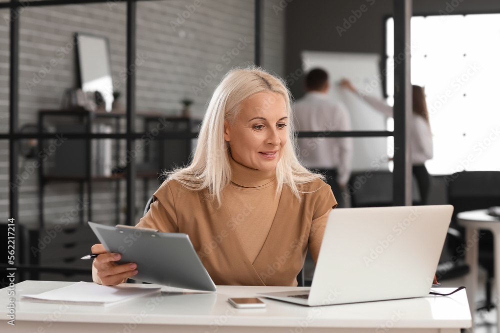 Mature bank manager working with laptop and clipboard in office