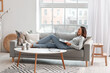 © Pixel-Shot - Young woman using laptop on grey couch in living room