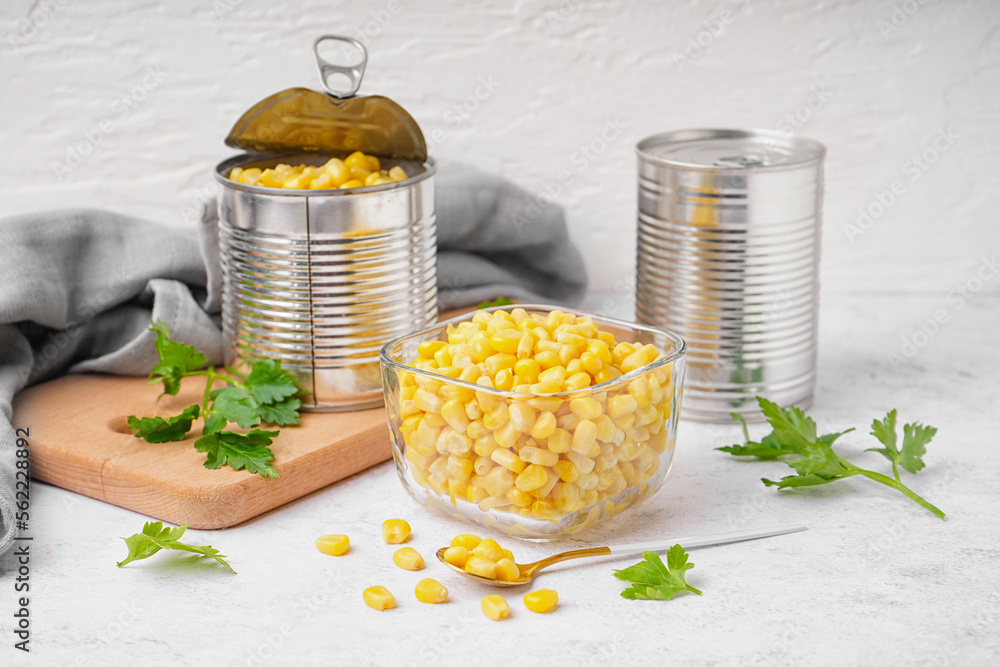 Bowl with corn kernels, tin cans, spoon and parsley on white table