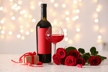 Glass Of Wine, Rose Flowers, Bottle And Gift On White Table Against Blurred Lights. Valentine's Day Celebration