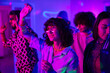 © pressmaster - Youthful brunette girl in white blouse and eyeglasses dancing among friends at home party in living room lit by neon light