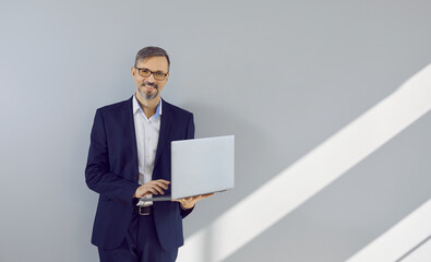 Wall Mural - Portrait of successful businessman with laptop. Happy mature business man in stylish dark blue suit, white shirt and eyeglasses standing by light grey copy space wall, holding computer and smiling