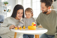 Family And Parenthood. Baby Girl And Her Caring Young Mom And Dad Are Playing With Rubber Toy Ducks Together. Young Caucasian Parents And Their Two-year-old Daughter Spend Time Together At Home.