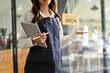 © bongkarn - Friendly Asian female waitress or coffee shop staff with her tablet, stands at the entrance door