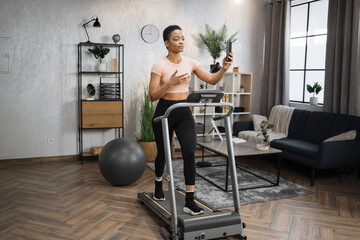 Wall Mural - Healthy young sports woman using phone having video conference while working out, running, doing cardio training on treadmill in morning time, indoor on background of modern apartment