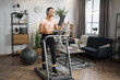 © Sofiia Shunkina - Full length photo of young active african business woman in sportswear and headset training at home working with tablet, doing cardio exercise on treadmill. Concept of sport, business.