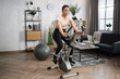 © Sofiia Shunkina - Front view of healthy female athlete having cardio workout indoors. Young healthy african woman training on stationary bike on the background of modern apartment.