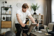 © sofiko14 - Home fitness workout, young caucasian man athlete training on smart stationary bike indoors watching on screen connected online to live streaming subscription service for biking exercise.