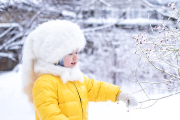 child playing with snowi n the park. smiling little girl in yellow jumpsuit and fluffy white hat walking on snowy winter day 