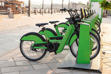 Electric Bicycles For Rent Are Green In Parking Lot In The City Center On The Street. Eco-friendly Mode Of Transport