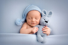A Cute Newborn Boy In The First Days Of Life Sleeps Naked On A Blue Fabric Background. The Kid Gently Hugs A Light Blue Knitted Bunny. Studio Professional Macro Photography, Newborn Baby Portrait.
