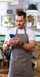 © ihorvsn - Portrait of happy young handsome Caucasian guy texting on smartphone standing in coffee house in apron at work, looking at camera and smiling. Restaurant service, business concept. Vertical video