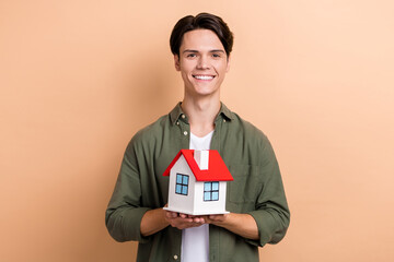 Poster - Portrait of satisfied cool cute man with brunet haircut dressed khaki shirt holding small house isolated on beige color background