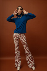 Wall Mural - Fashionable confident woman wearing trendy brown rectangular sunglasses, stylish blue turtleneck, flared trousers with zebra print, boots. Full-length studio portrait