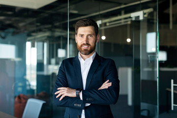 Portrait of confident smiling middle aged businessman in formal wear posing with crossed hands in office interior