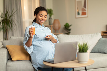 Positive young pregnant black woman video calling to doctor and showing medical pills bottle to laptop, sitting on couch