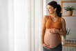 © Dorde - Happy pregnant woman standing by the window, wearing an orange sports bra and gray leggings is smiling and touching her stomach.