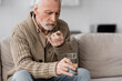 © LIGHTFIELD STUDIOS - senior man with parkinsonian syndrome sitting on couch and holding pill and glass of water in trembling hands.