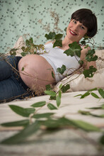 Portrait Of Pregnant Woman Wrapped With Branch Of Green Leaves While Lying On Bed