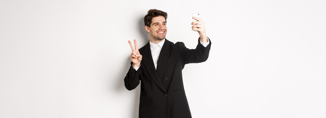 Portrait of good-looking man taking selfie on new year party, wearing suit, taking photo on smartphone and showing peace sign, standing against white background