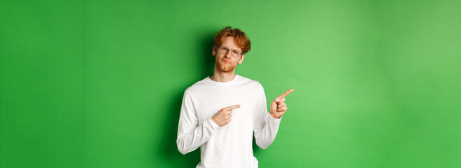 Wall Mural - Doubtful redhead guy in glasses pointing fingers left, looking hesitant and unsure at camera, standing over green background