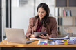 © David - Freelancer Happy business Asian woman in knitwear taking notes at laptop sitting at desk office, finance concept.