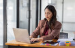 © David - Freelancer Happy business Asian woman in knitwear taking notes at laptop sitting at desk office, finance concept.