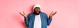 Confused african-american man in beanie looking at upper left corner doubtful, shrugging uncertain, standing over pink background