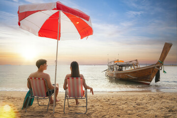 Asian traveller couple sitting and see sunset togather on the beach with longtail boat background