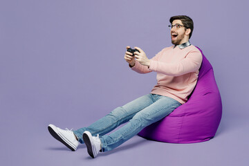 Wall Mural - Full body fun young IT man wear casual clothes pink sweater glasses sit in bag chair hold in hand play pc game with joystick console isolated on plain pastel light purple background studio portrait.