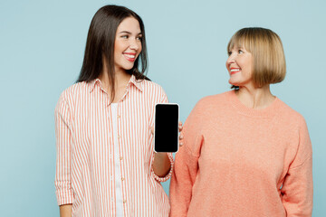 Wall Mural - Fun elder parent mom with young adult daughter two women together wear casual clothes hold in hand use mobile cell phone with blank screen area isolated on plain blue background. Family day concept.