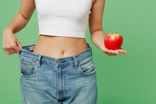 Cropped Young Woman Wears White Clothes Show Loose Pants On Waist After Weightloss Hold Red Apple Isolated On Plain Light Green Background. Proper Nutrition Healthy Fast Food Unhealthy Choice Concept.