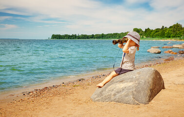 Wall Mural - Little girl sitting on the stone on the seashore with binocular in her hands.